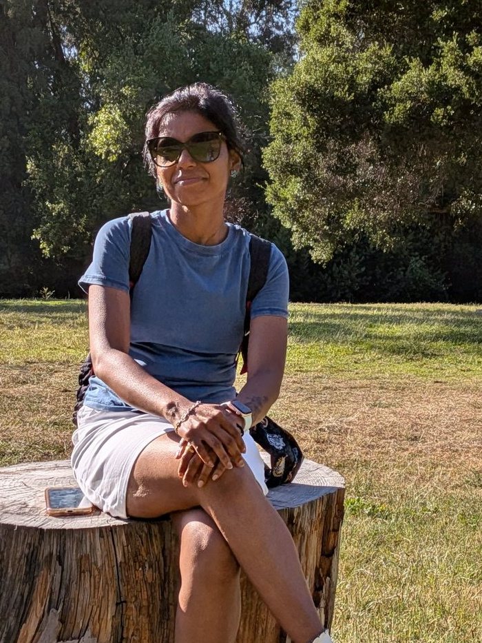 Nithya Sekar sitting on a tree stump in a sunny park, wearing a blue t-shirt, white shorts and sunglasses